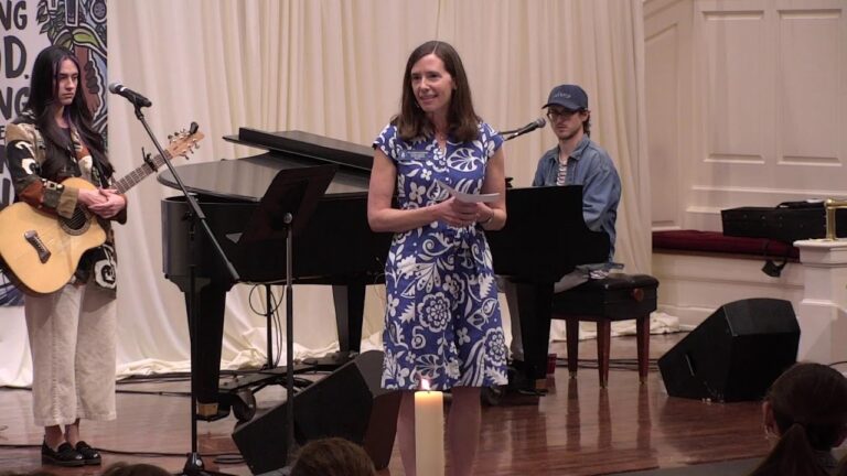 Woman in a blue floral dress speaks on stage with a pianist and guitarist beside her; a candle sits at the front on the floor.