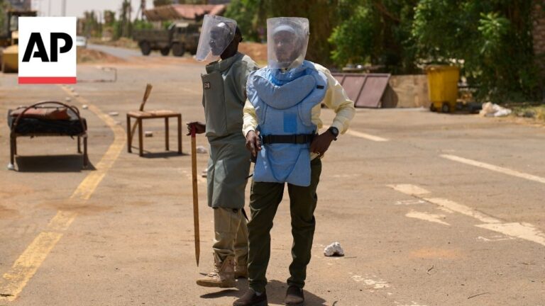 Two people in hazmat suits with face shields stand on a dusty street with debris; AP watermark in the top left corner.