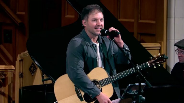 Male musician singing into a handheld mic while playing an acoustic guitar on stage with a grand piano in the background, wearing a dark jacket and white shirt.