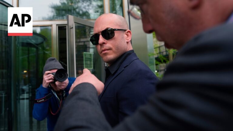 Bald man in a dark suit and sunglasses stands outside a glass entry as a photographer films him nearby (AP logo visible).