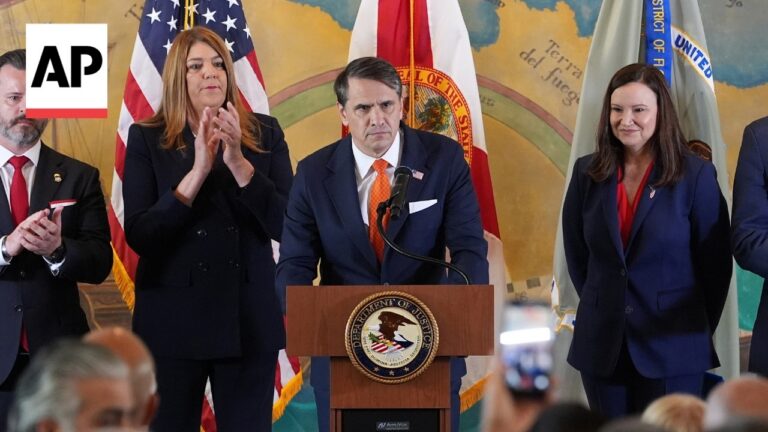 Man in a navy suit and orange tie speaks at a DOJ podium, flanked by officials with flags behind; AP News logo in the corner.