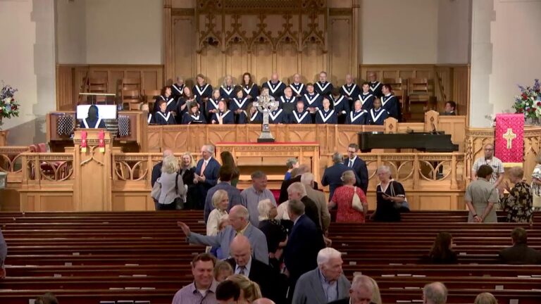 Choir in black robes with white collars standing on a raised chancel in a church, congregation mingling in the pews.