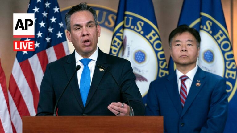 AP Live logo on the left; a man in a dark suit speaks at a wooden podium with microphones, USA flag and Capitol banners behind him, another man in a blue suit stands beside.