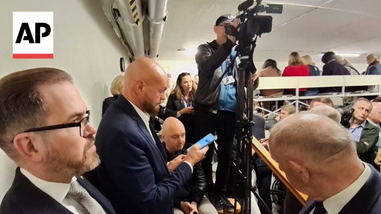 AP logo in the top-left corner; a man with glasses in a suit on the left, another bald man in a suit looking at his phone, and a cameraman with a tripod amid a crowded stairwell of journalists and onlookers.