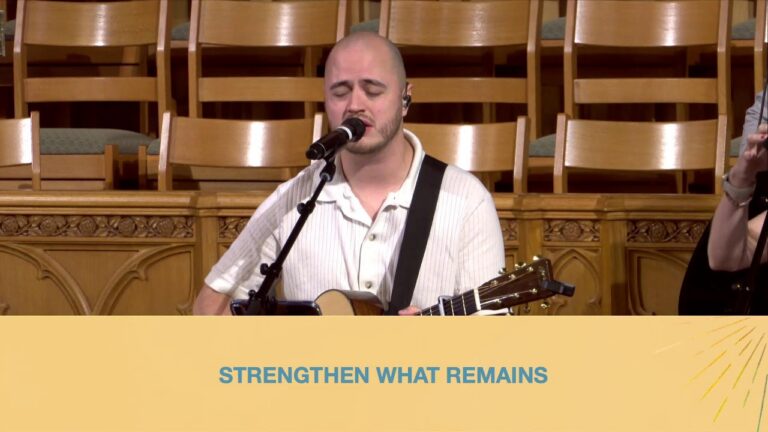Musician with a guitar singing into a microphone on a wooden stage, banner at bottom reads 'STRENGTHEN WHAT REMAINS'.