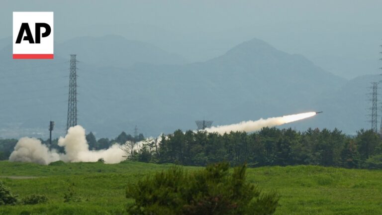 Missile launches from a ground site, leaving flame and smoke across a grassy field with trees and distant mountains; AP logo in the upper left corner.