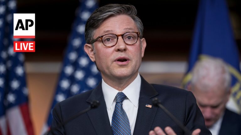 Man in a dark suit and glasses speaks at a press conference with the AP Live logo in the corner and American flags in the background.