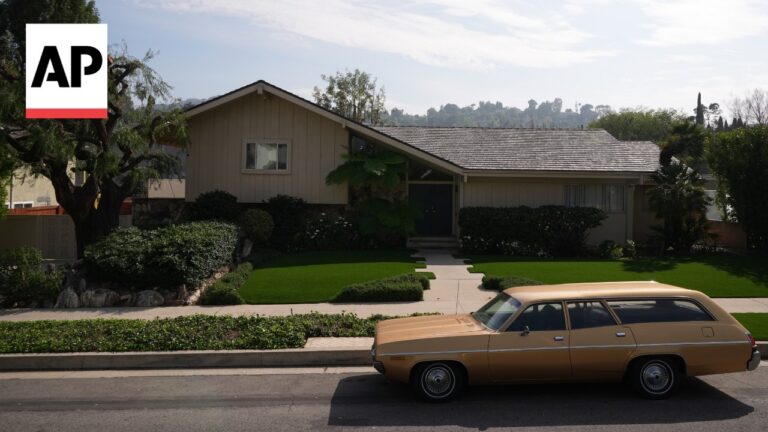 Mid-century suburban home with a manicured lawn and a tan vintage wagon parked on the street; AP logo in the top-left corner.
