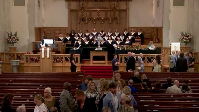 Choir on stage in a church sanctuary with floral arrangements as attendees mingle in pews.