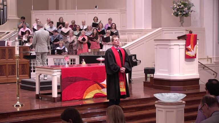 Choir singing on risers behind a conductor in a gray suit at the front of a church service.