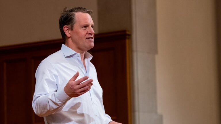 Man in a white button-down shirt speaks to an audience, gesturing with his right hand in a lecture setting