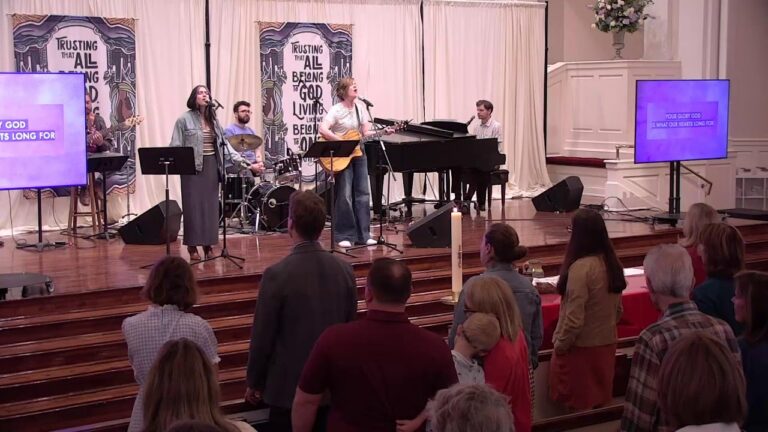 Worship band performing on a church stage with singer, guitarist, drummer, and pianist while the congregation stands nearby.