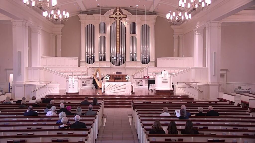 Church sanctuary with a large cross and pipe organ above the altar; attendees sit in pews facing the front.
