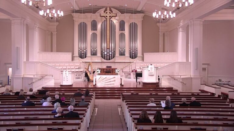 Church sanctuary with a large cross and pipe organ above the altar; attendees sit in pews facing the front.