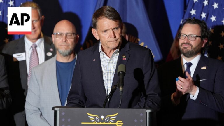 Man in a navy blazer speaks at a podium with an AP logo, flanked by supporters, with American flags in the background.