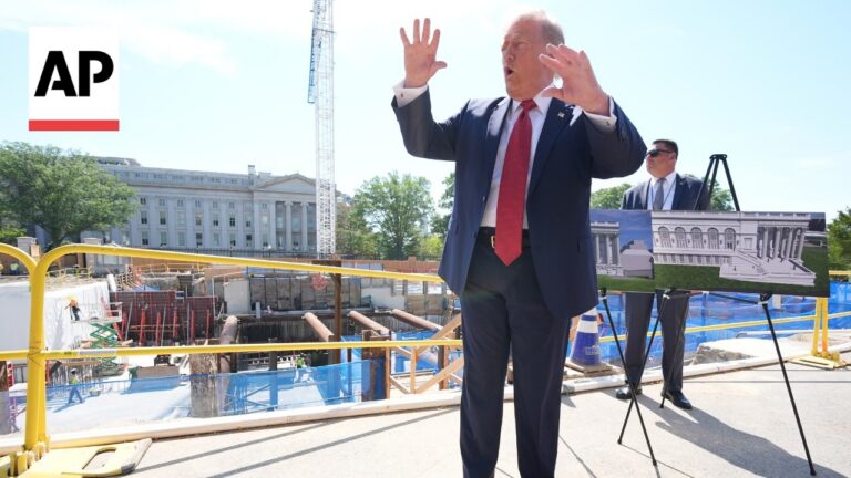 Man in a dark suit and red tie speaks and gestures at a construction site, with an architectural rendering on an easel and the AP logo in view.