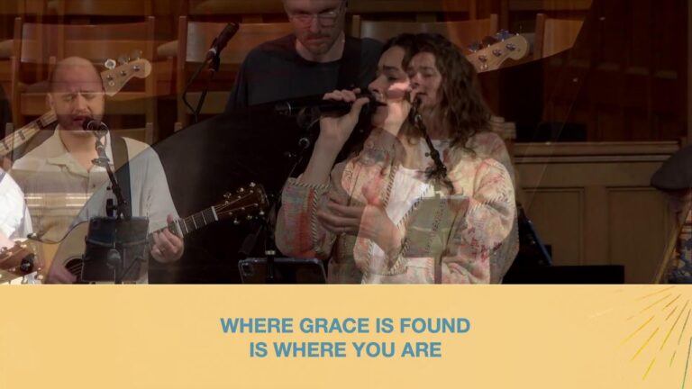 Worship band performing on stage: a female singer center, guitarist left, banner reads 'Where grace is found is where you are'.