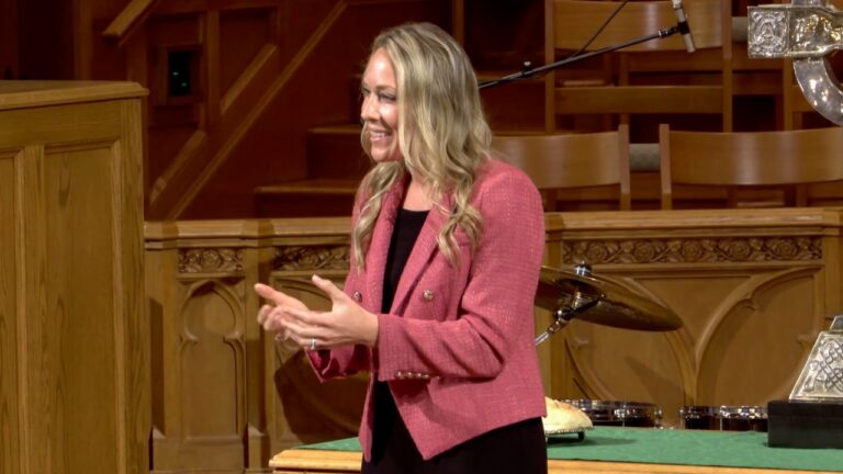 Woman in a pink blazer speaking at a church podium with hands gesturing.