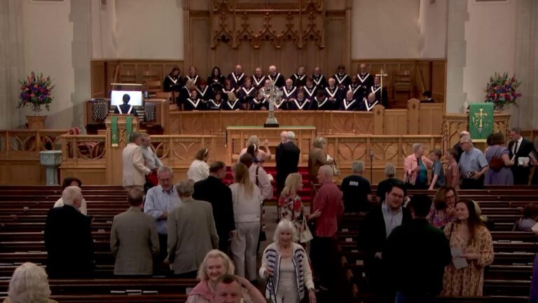 Choir in robes stands on a raised chancel in a church, with attendees mingling in the nave below and an altar in front.