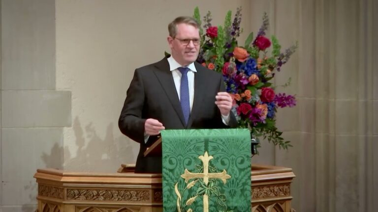 Man in a suit delivering a sermon at a wooden pulpit with a green banner featuring a gold cross, in front of a colorful floral arrangement.