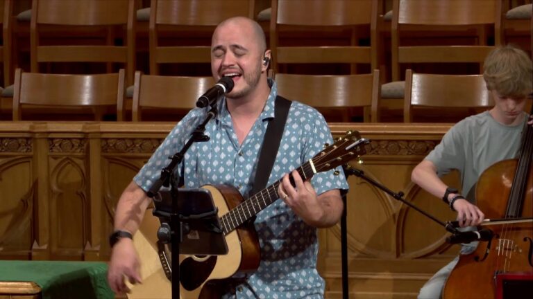 Male singer with an acoustic guitar performing on a wooden stage, eyes closed, microphone in front.