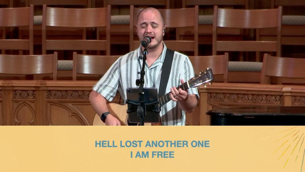 Male singer with an acoustic guitar performs on a wooden stage; a yellow banner below displays the lyric 'HELL LOST ANOTHER ONE I AM FREE'.