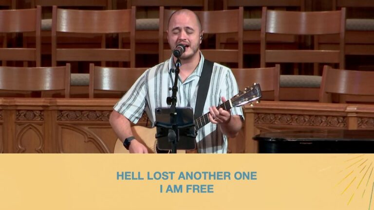Male singer with an acoustic guitar performs on a wooden stage; a yellow banner below displays the lyric 'HELL LOST ANOTHER ONE I AM FREE'.