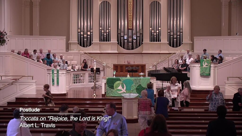 Church sanctuary with a large organ in back, audience seated and standing near the front, and green banners; on-screen caption reads "Postlude Toccata on 'Rejoice, the Lord is King!'" by Albert L. Travis