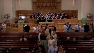 Choir in matching robes stands on a wooden platform behind a raised altar inside a church.