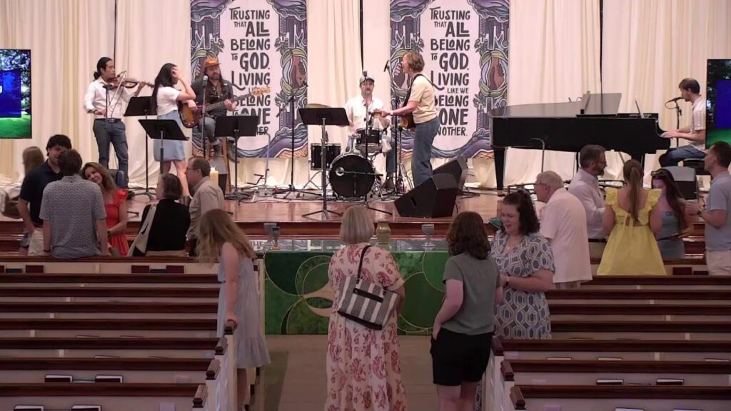 Church band performing on stage with violin, guitar, drums, and piano as attendees chat in the pews.
