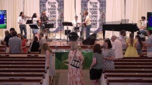 Church band performing on stage with violin, guitar, drums, and piano as attendees chat in the pews.