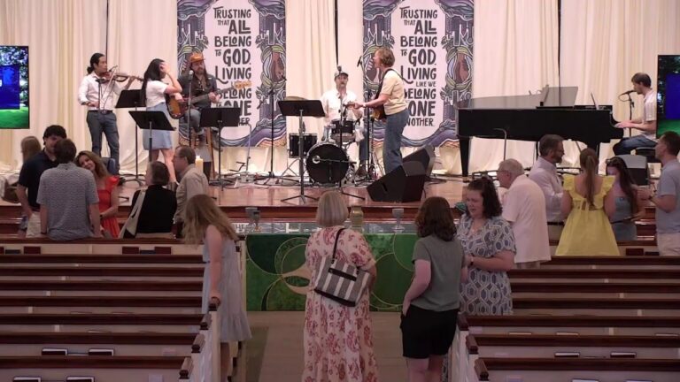 Church band performing on stage with violin, guitar, drums, and piano as attendees chat in the pews.
