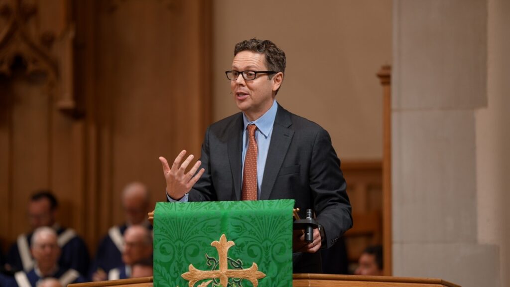 Man in a suit giving a speech at a lectern draped with a green cloth featuring a gold cross in a church setting.