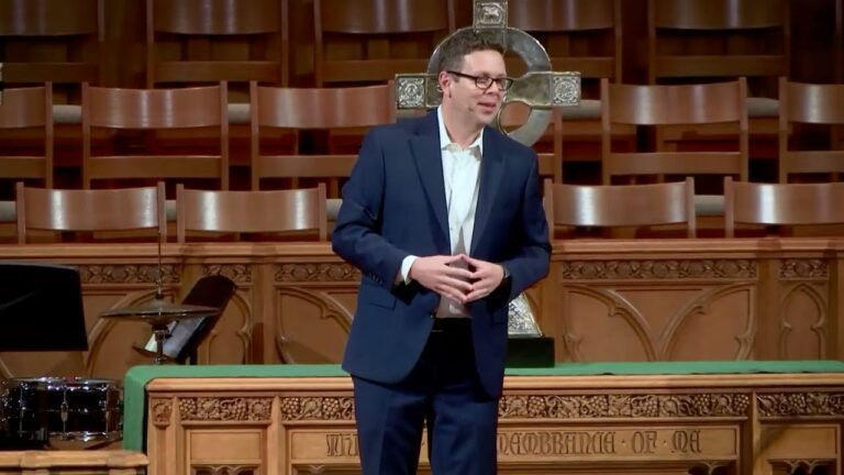 A man in a navy suit speaks from an ornate church pulpit with a large cross behind him and empty wooden chairs in the background.