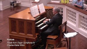 Man in a suit playing a wooden pipe organ, with sheet music on the stand, in a recital space with audience seating nearby.