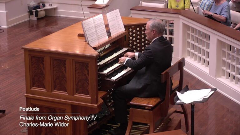 Man in a suit playing a wooden pipe organ, with sheet music on the stand, in a recital space with audience seating nearby.