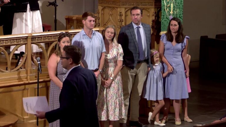 Group of adults and two children lined up on a church stage, a man in the foreground hands out papers near a microphone. There is ornate wood railing behind them and a green banner to the side.