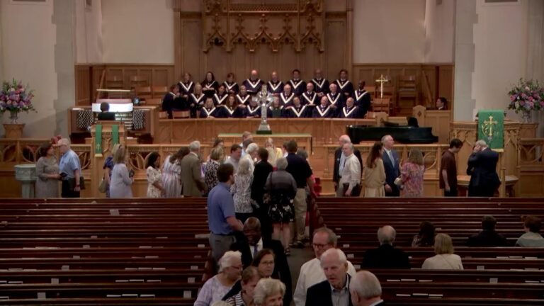Choir in black robes with white collars stands on risers behind the altar as guests mingle in a large church sanctuary.