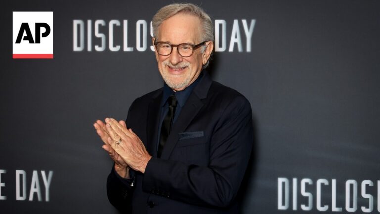 Smiling older man in a dark suit claps on a backdrop with the AP logo and the words 'DISCLOSURE DAY' behind him.