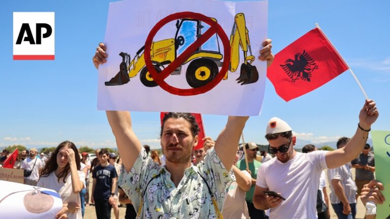 Crowd of protesters outdoors; man in foreground raises a sign with a crossed-out excavator image, AP logo at left, Albanian flag waving nearby.