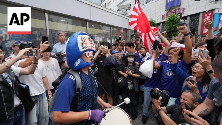AP logo on the left; a drummer wearing a blue mask and jersey plays a drum as a large crowd films and cheers nearby.