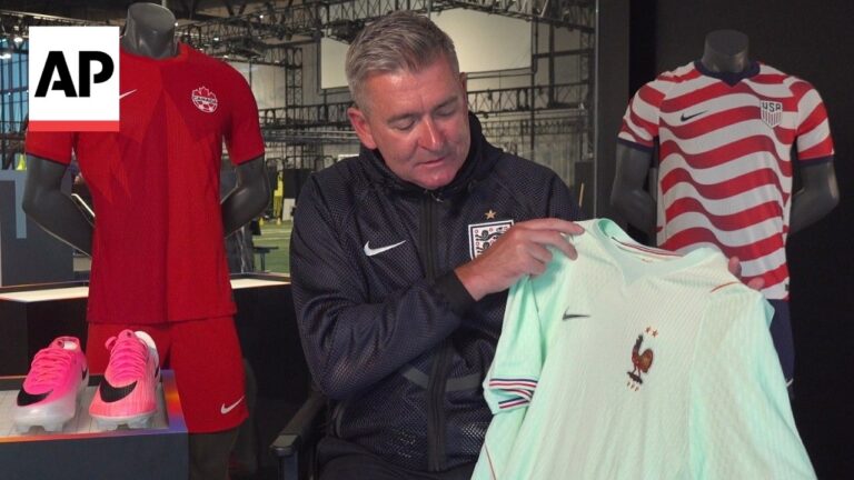 Man in a dark jacket signs an England football shirt in a showroom, with display mannequins in red Canada kit and USA striped jersey in the background.