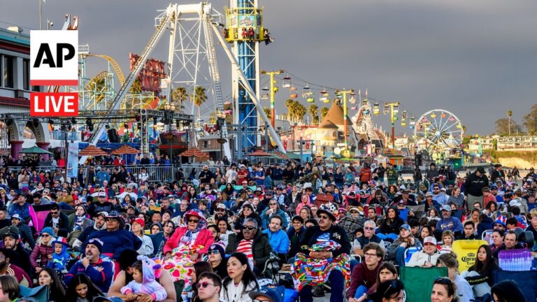 Large crowd seated on folding chairs at a seaside amusement park, with rides and a ferris wheel in the background.