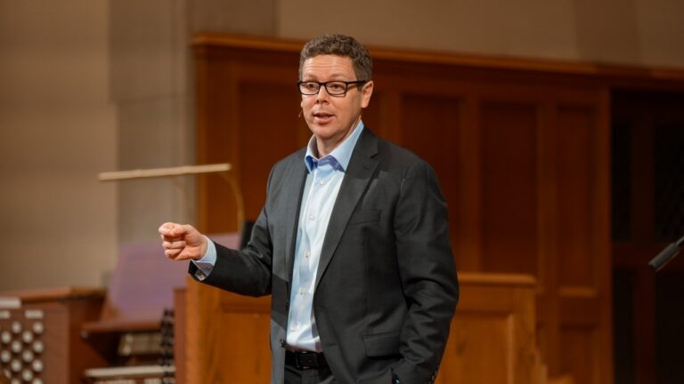 Man in a dark suit and glasses giving a lecture on a wooden stage with warm wood panels behind him.