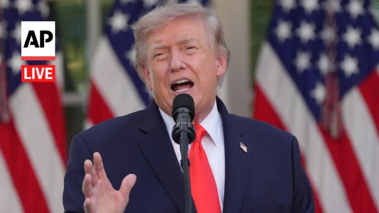 Man in a dark suit and red tie speaks into a microphone with American flags behind; AP Live logo left.