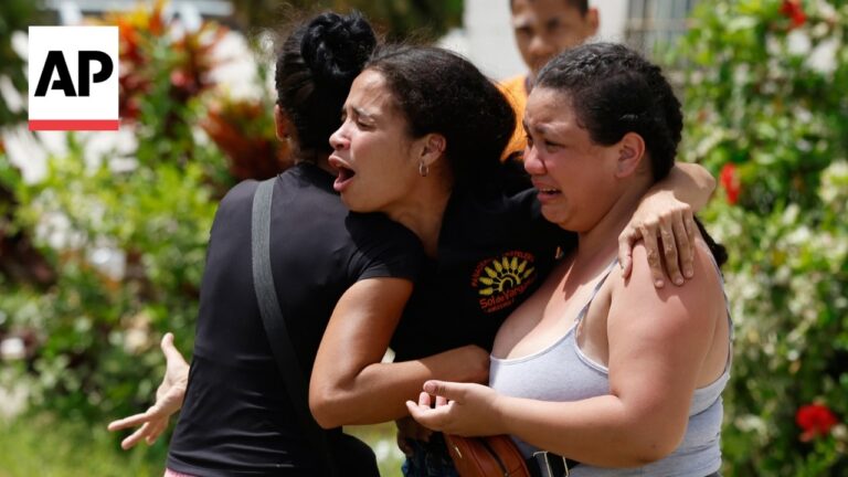 Two women hug and cry outdoors, one in a gray tank top and the other in black, with an AP logo in the corner in the frame.