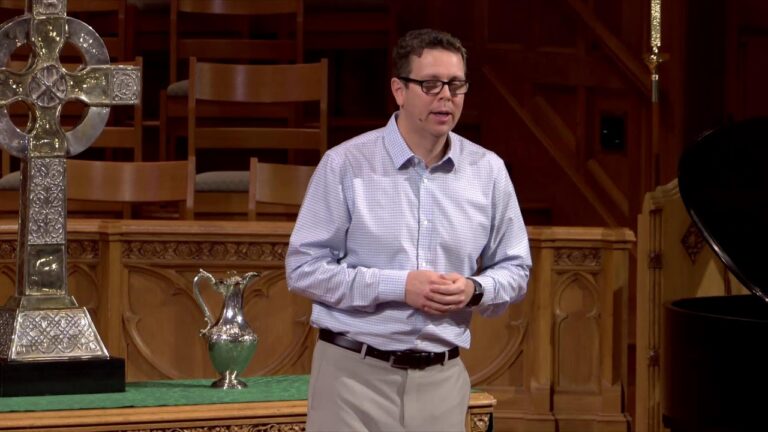 Man in a light blue check shirt and glasses speaks in a church, hands clasped, with a large silver cross and pitcher on the table beside him