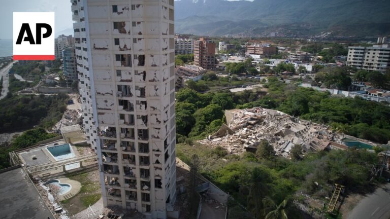 AP photograph: a tall, damaged apartment building with broken windows and exposed interiors, rubble in a nearby lot, with a city and mountains in the background.