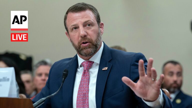 Man in a blue suit testifying at a hearing, speaking into a microphone with an AP Live logo nearby and his hand raised for emphasis.