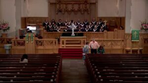 Church sanctuary with a choir on the balcony behind the altar, robes in black with white V-shaped collars, and a central decorative cross; three people in the aisle near the front interact near the stage.
